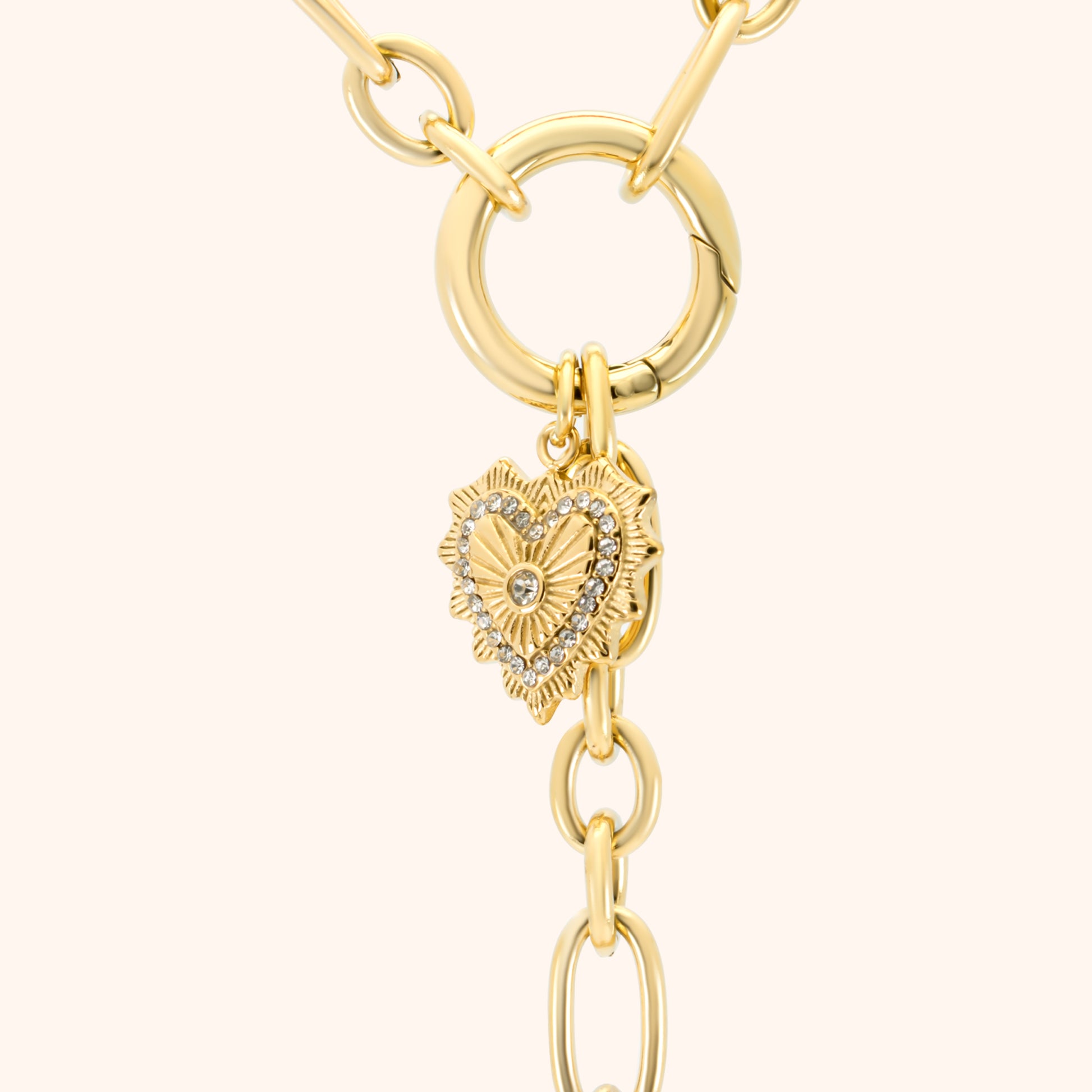 Close-up of the pavé heart charm on the PRESSIE Radiant Heart Lariat necklace; 18k gold plated with radiating texture and clear stone details, connected to a large circular clasp.
