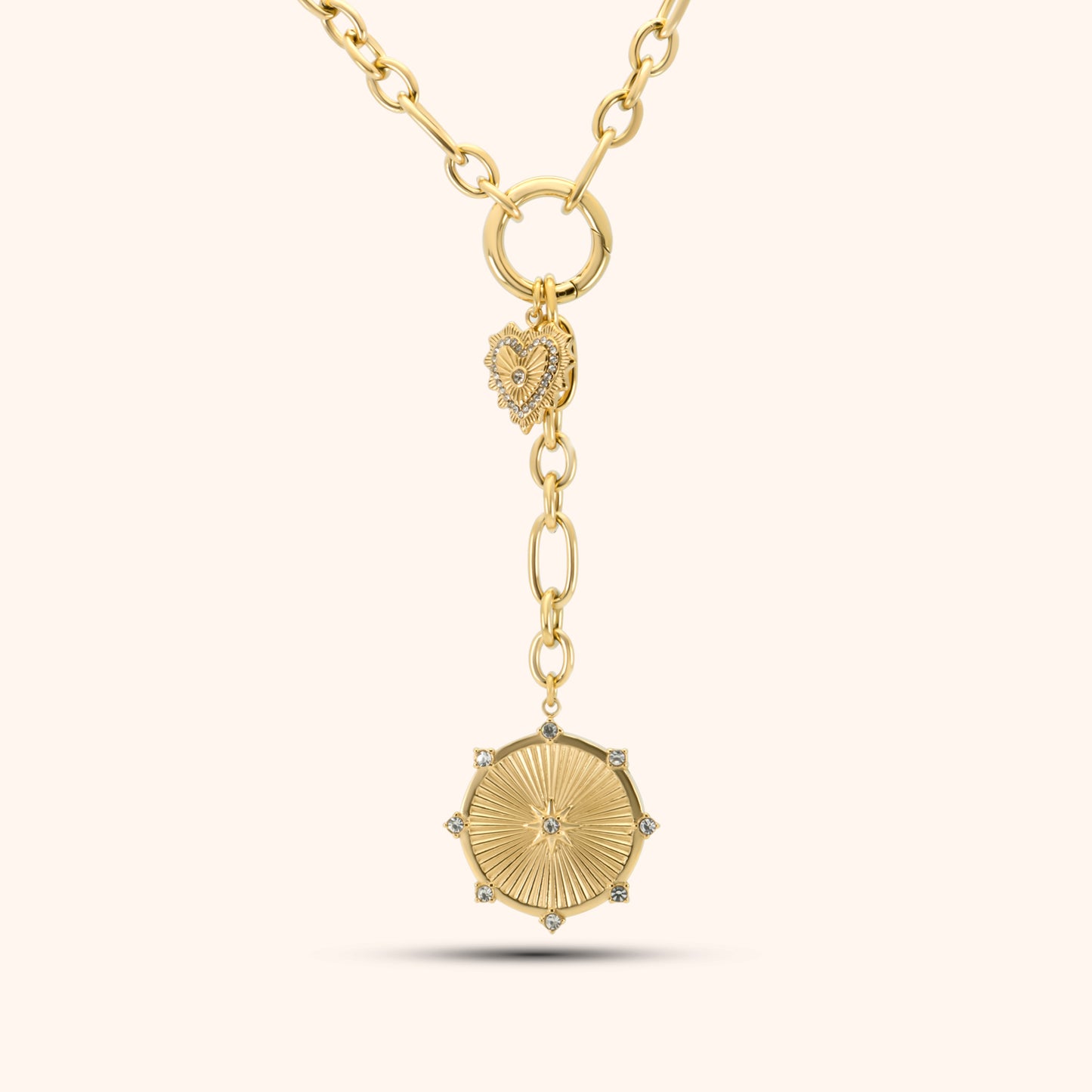 PRESSIE Radiant Heart Lariat necklace in 18k gold plate; features mixed paperclip chain, a central circular connector with a pavé heart charm, and a lariat drop ending in a sunburst medallion with clear stone accents.